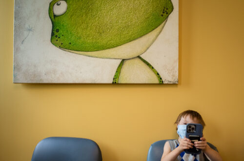 color photograph of boy in blue chair against yellow wall under green frog taking a video of photographer in a doctor's waiting room in Little Rock, Arkansas, that is part of the story we tell project.