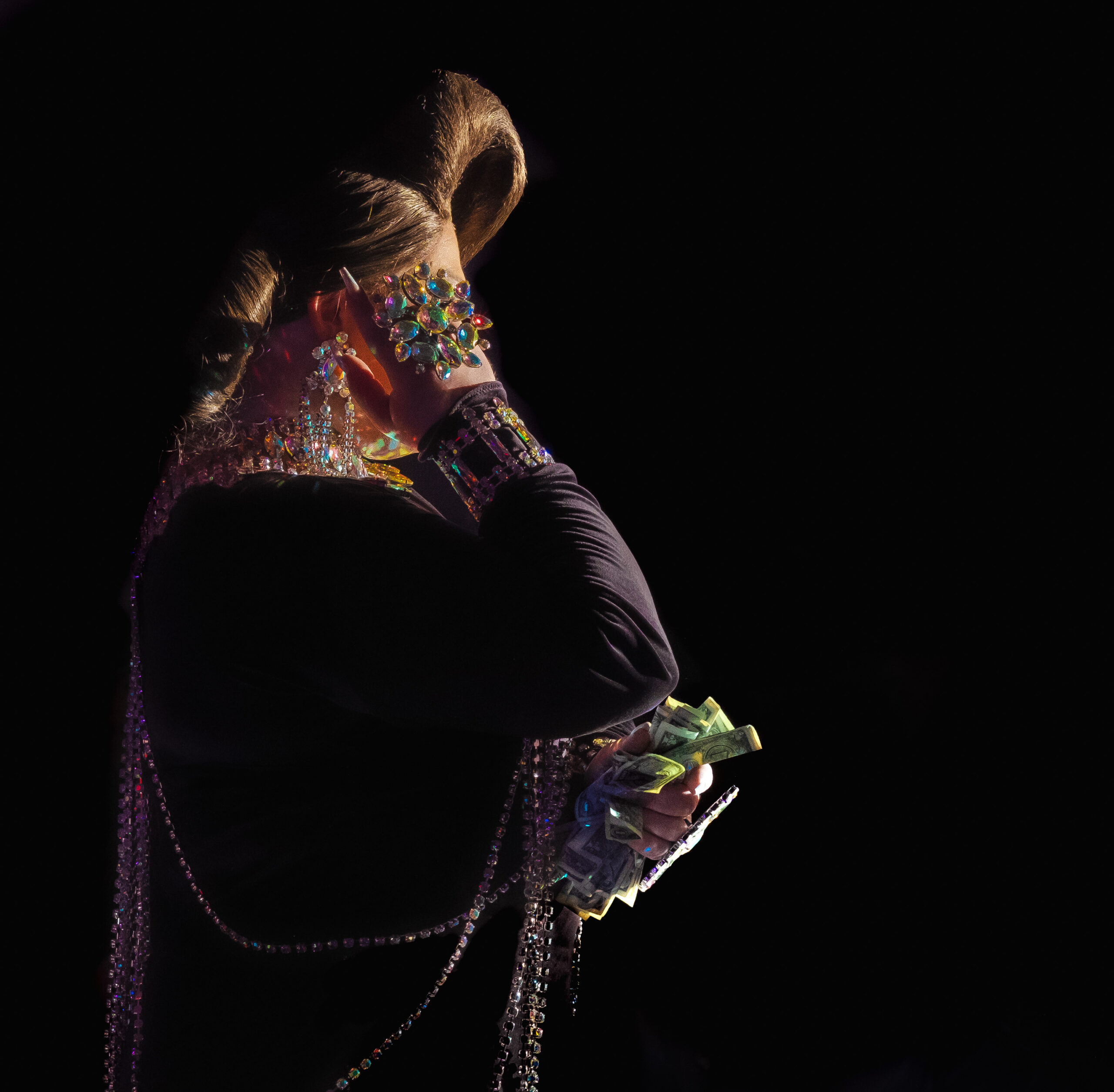 Color photograph taken by Chris Attig Blue Bus Photos showing Arkansas Drag Queen Melanie Masters performing drag during an entertainment intermission at the 2023 Miss Gay Central Arkansas America Drag Pageant in Conway, Arkansas, while carrying dollar bills in one hand and wearing reflective jewelry on the other hand.