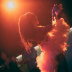 color image of Little Rock Arkansas Drag Queen Aubi Gold performing in a yellow top and white feathered wings at Diamonds and Pearls at Club Sway in orange and red tone casts