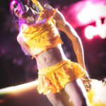 color image of Little Rock Arkansas Drag Queen Aubi Gold performing in a yellow top and white feathered wings at Diamonds and Pearls at Club Sway in a yellow skirt and top with the pink neon sway sign in the background