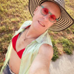 little rock arkansas trans woman queer photographer Gynger St Claire poses for a saelfie wearing blum denim shorts, a red bikini, a green and white checkered coverup and a black and white striped sunhat