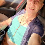 little rock arkansas trans woman queer photographer Gynger St Clair poses for a seated selfie in her car wearing pink tinted glasses, a sky blue cropped tank top and navy blue sheer bolero with a dark tanned pleather skirt