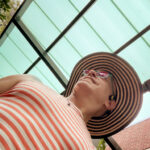 Trans woman and queer photographer Gynger St Claire takes a selfie while walking under an awual blue awning wearing annorange and white striped cropped tank top