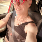 little rock arkansas trans woman queer photographer Gynger St Clair poses for a seated selfie in her car wearing red glasses that evoke Iris Apfel, a black cropped tank top and green denim shorts