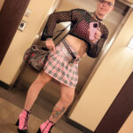 little rock arkansas trans woman queer photographer Gynger St Clair poses for a full length mirror shot wearing pink combat boots with 4" heels, a plaid pink and black mini skirt, a pink and black bb belt, a mesh top over a cami with a shelf bra and a spiked dog collar with an owl charm hanging from the center