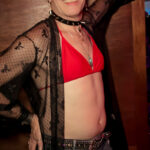 little rock arkansas trans woman queer photographer Gynger St Clair poses for a selfie at Club Sway during Pride 2025, we aring a red bikini top, blue denim shorts and a black sheer coverup, as well as a dog collar with spikes and a dark red heart pendant hanging from the collar